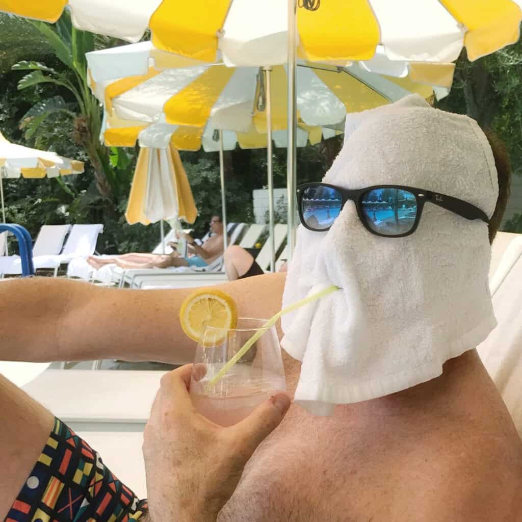 Man with a towel over his face, wearing sunglasses and pretending to sip a cocktail.