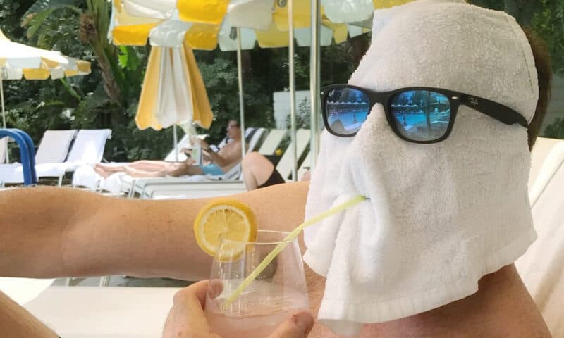 Man with a towel over his face, wearing sunglasses and pretending to sip a cocktail.