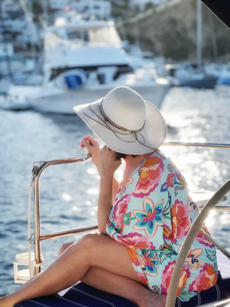 Girl in colorful dress and hat sitting on a sailboat, looking away towards the sunset.
