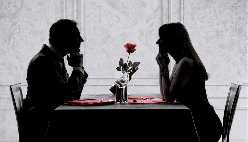 A silhouette of a couple dining out. A rose sits on the table between them.