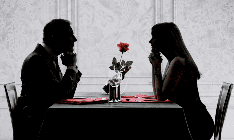 A silhouette of a couple dining out. A rose sits on the table between them.