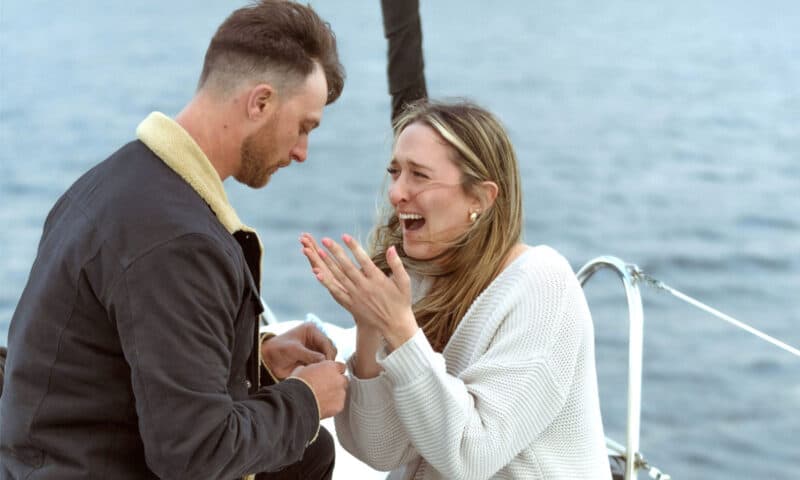 A man on one knee, proposing to his girlfriend on the deck of the Riviera sailboat in the San Diego Bay.