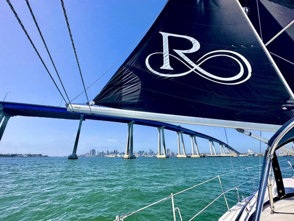 The navy blue and white sail of the SV Riviera with the water and Coronado Bridge in the background. 