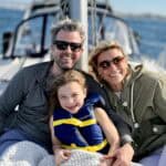 A mother, father, and daughter in a life jacket on the deck of a yacht at sail.