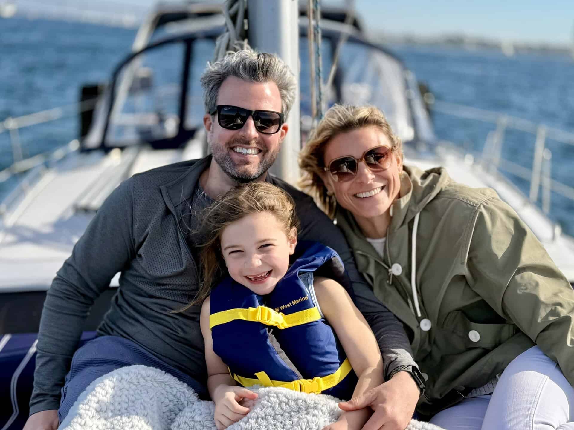 A mother, father, and daughter in a life jacket on the deck of a yacht at sail.