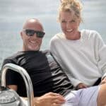 A couple poses on the deck of a sail boat, with the ocean behind them