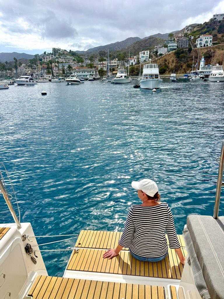 A woman sits on the edge of a Rivera sailboat deck in the Avalon harbor, with the city in the background.