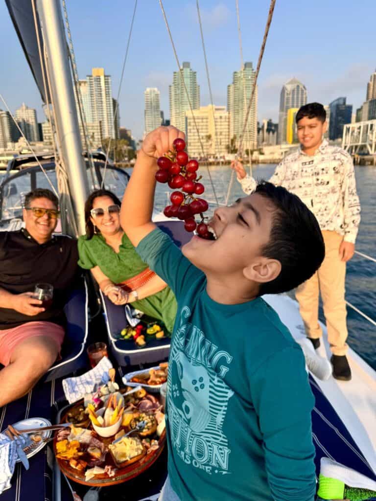 Boy on a boat enjoying grapes with friends and a scenic harbor backdrop.