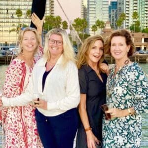 Four women holding cocktails, posing for the camera on the deck of a sailboat, with downtown San Diego in the background.